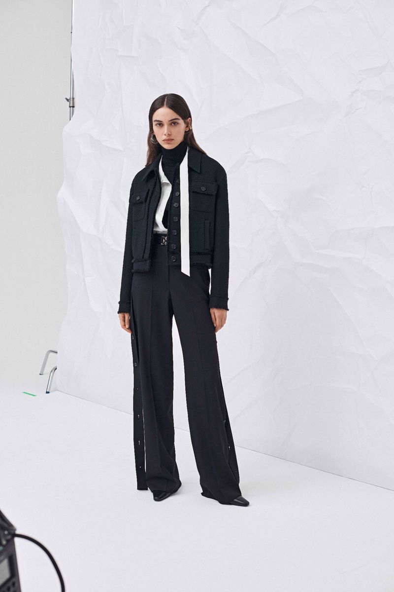 Studio shot of a woman in a black jacket and wide-leg pants against a white backdrop, modelling fashion, and wearing a turtleneck sweater.
