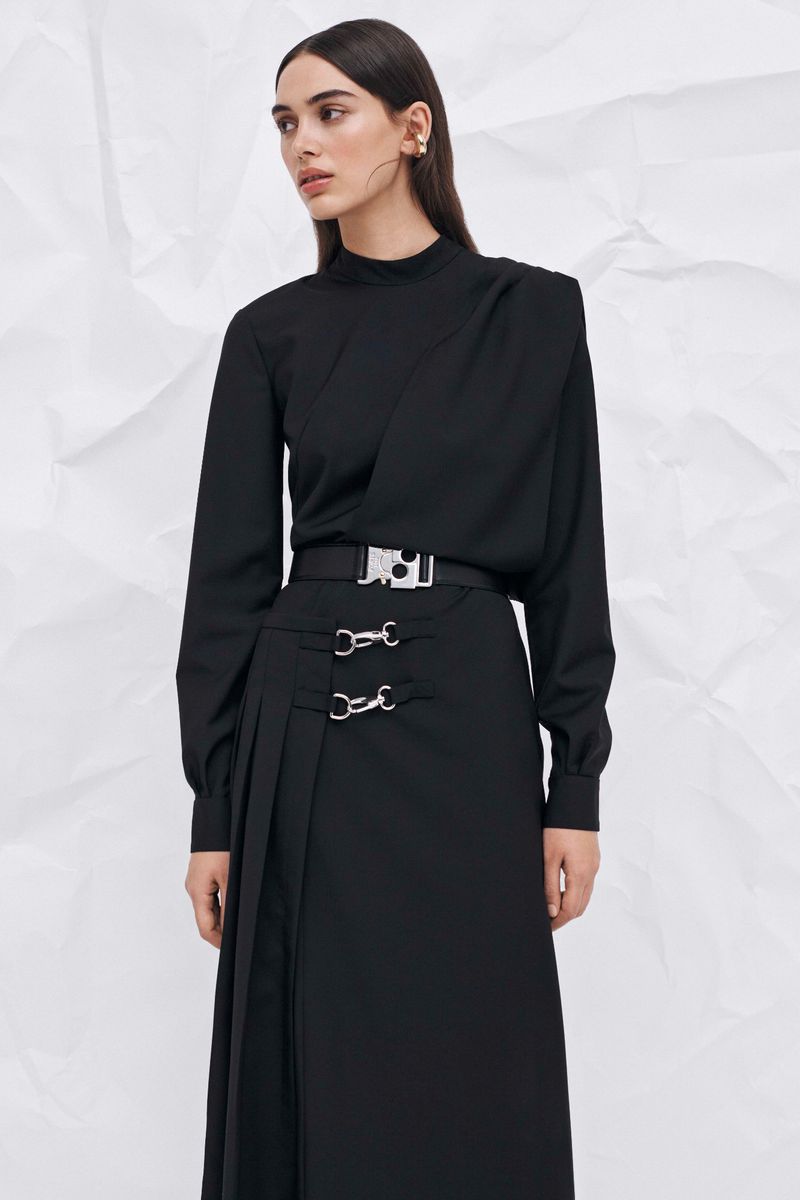 A woman poses indoors in a black long-sleeve top and a long black skirt with silver hardware against a white wrinkled backdrop.