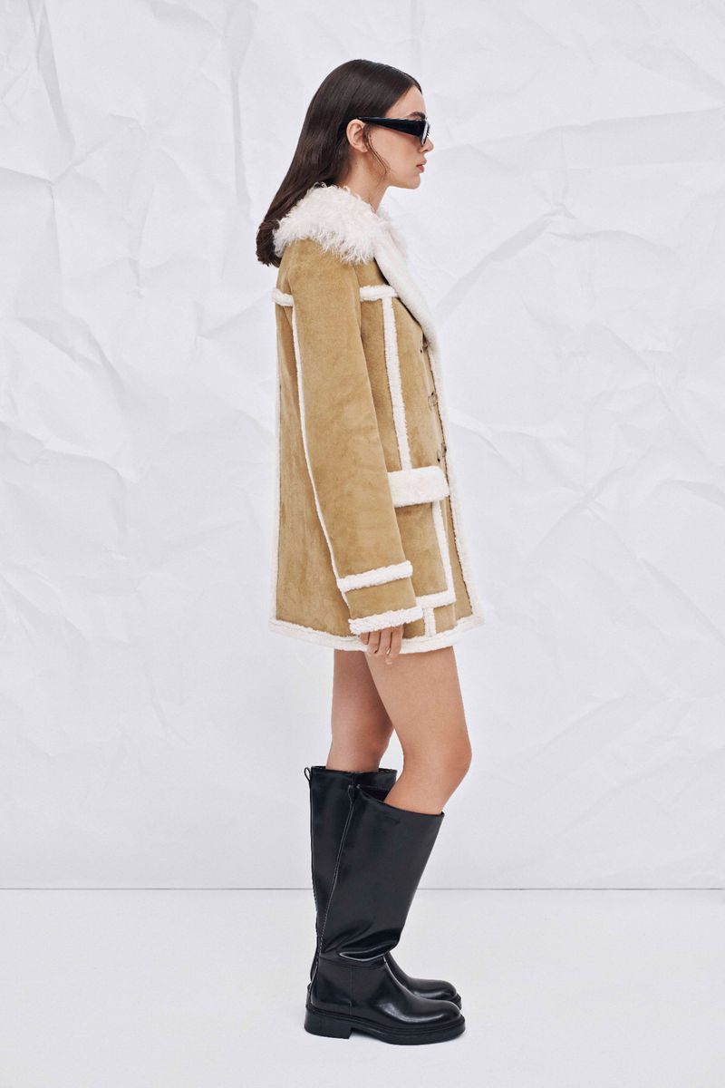 A woman in a tan coat with white trim and black boots poses in front of a white backdrop in a fashion editorial shot.
