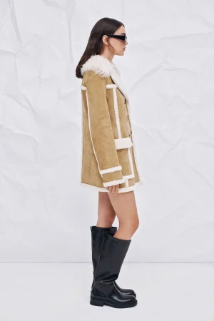 A woman in a tan coat with white trim and black boots poses in front of a white backdrop in a fashion editorial shot.