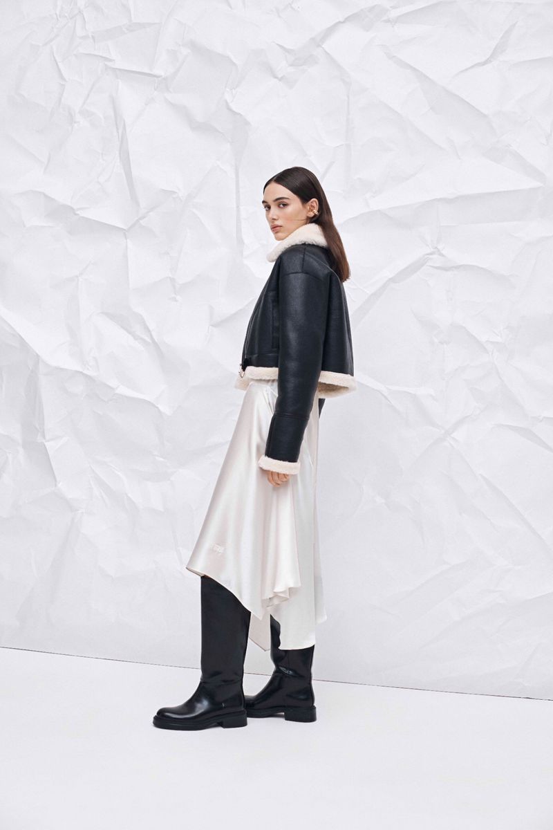 Fashionable woman in a shearling-lined jacket and flowing skirt poses against a white wrinkled backdrop in a studio environment.