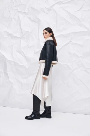 Fashionable woman in a shearling-lined jacket and flowing skirt poses against a white wrinkled backdrop in a studio environment.