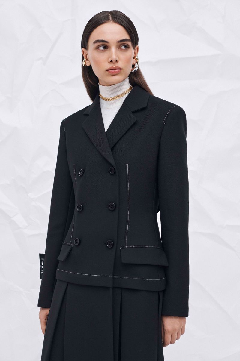 Portrait of a woman wearing a black double-breasted blazer with white stitching and a pleated skirt against a textured white background.