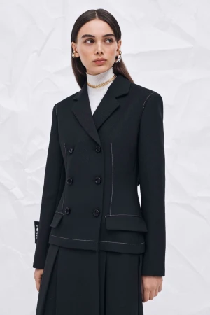 Portrait of a woman wearing a black double-breasted blazer with white stitching and a pleated skirt against a textured white background.