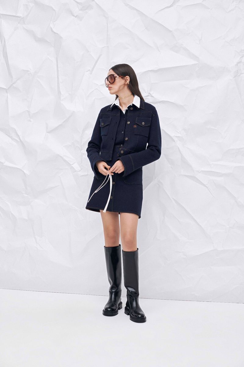 Full shot of a woman posing in a navy jacket, mini skirt and black boots in front of a white textured background in a studio.