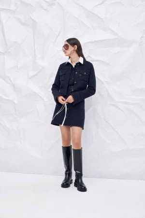 Full shot of a woman posing in a navy jacket, mini skirt and black boots in front of a white textured background in a studio.