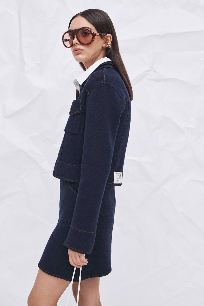 A fashionable adult female posing in a navy-blue jacket and skirt with oversized sunglasses against a textured white backdrop in a studio.