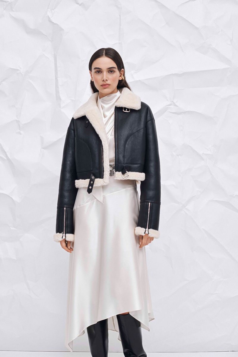 A woman models a black, cropped leather jacket with a white shearling collar and a long, white silk skirt against a crumpled paper backdrop.