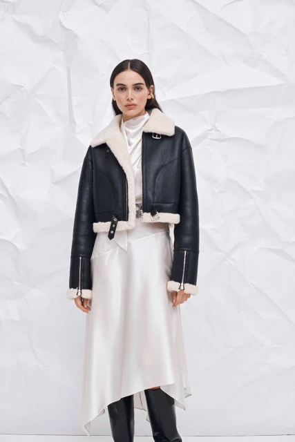 A woman models a black, cropped leather jacket with a white shearling collar and a long, white silk skirt against a crumpled paper backdrop.