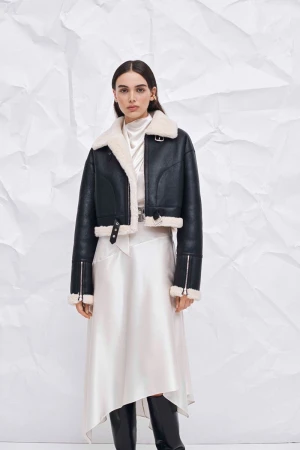 A woman models a black, cropped leather jacket with a white shearling collar and a long, white silk skirt against a crumpled paper backdrop.