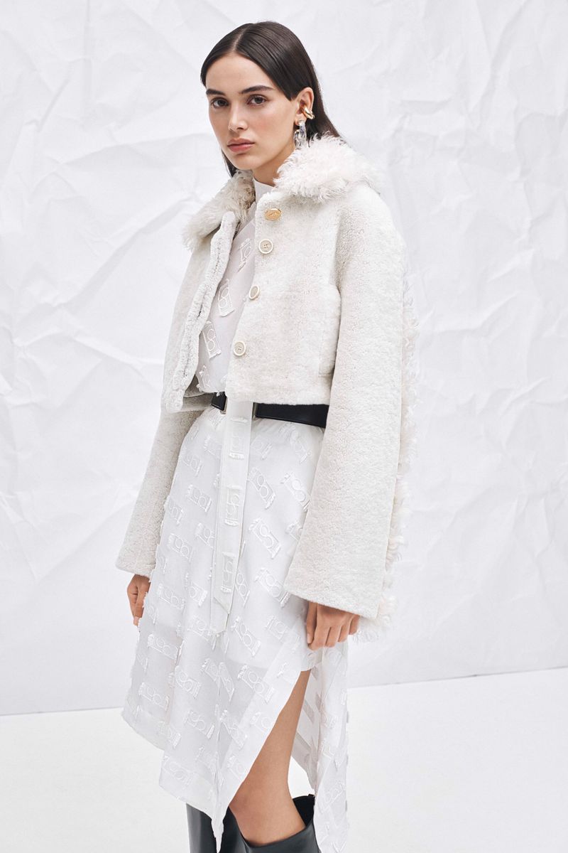 A woman stands in a bright studio wearing a cream-colored shearling jacket and a white dress with black boots, against a white background.