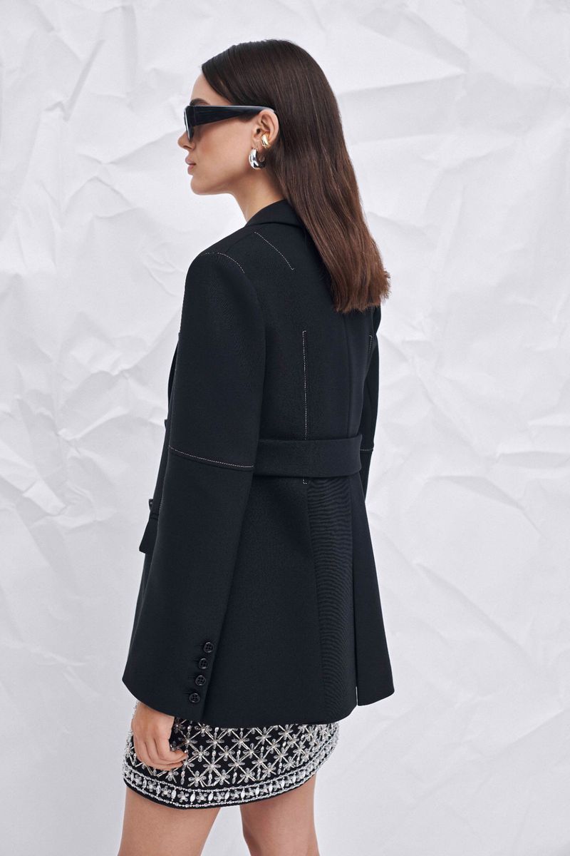 Profile of a woman wearing a black belted blazer, sunglasses, and a patterned skirt, against a textured white paper background.