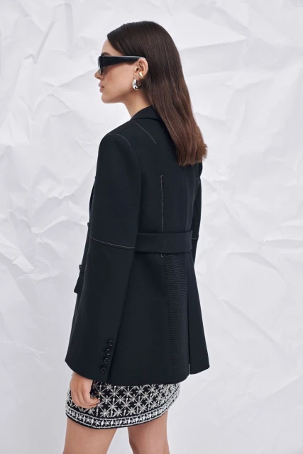 Profile of a woman wearing a black belted blazer, sunglasses, and a patterned skirt, against a textured white paper background.