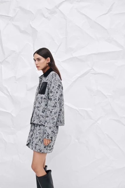 Woman in floral print jacket and skirt, paired with black boots, poses against a textured white backdrop in a fashion editorial shot.
