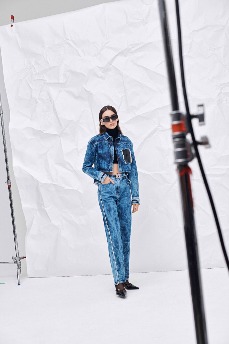 A woman poses in a denim jacket and jeans against a white backdrop in a fashion shot, with a blurred light stand in the foreground.