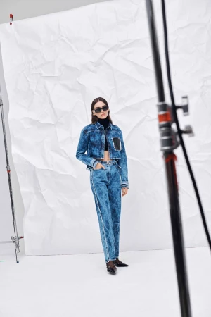 A woman poses in a denim jacket and jeans against a white backdrop in a fashion shot, with a blurred light stand in the foreground.