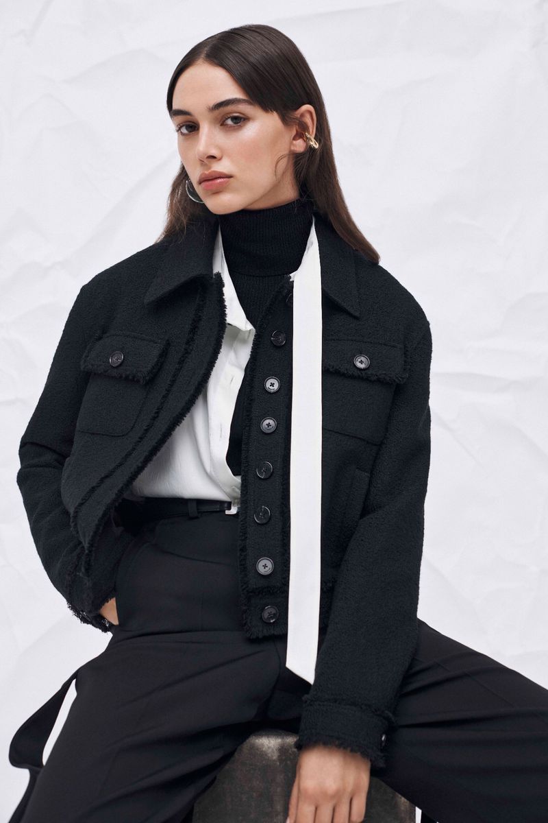 Stylish woman in a black tweed jacket, black turtleneck, wide-legged trousers, accessorized with hoop earrings and a white scarf.