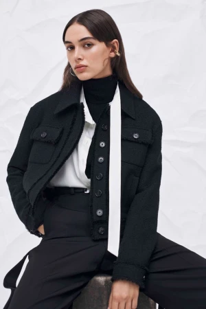 Stylish woman in a black tweed jacket, black turtleneck, wide-legged trousers, accessorized with hoop earrings and a white scarf.