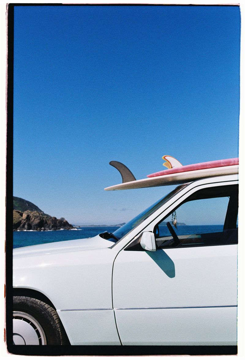 Surfboards secured on a vintage white car parked near a beach, with clear blue sky. A coastal landscape in the distance.