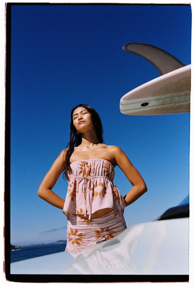 Woman posing in pink floral two-piece outfit with surfboard and clear blue sky backdrop in sunny outdoor setting.
