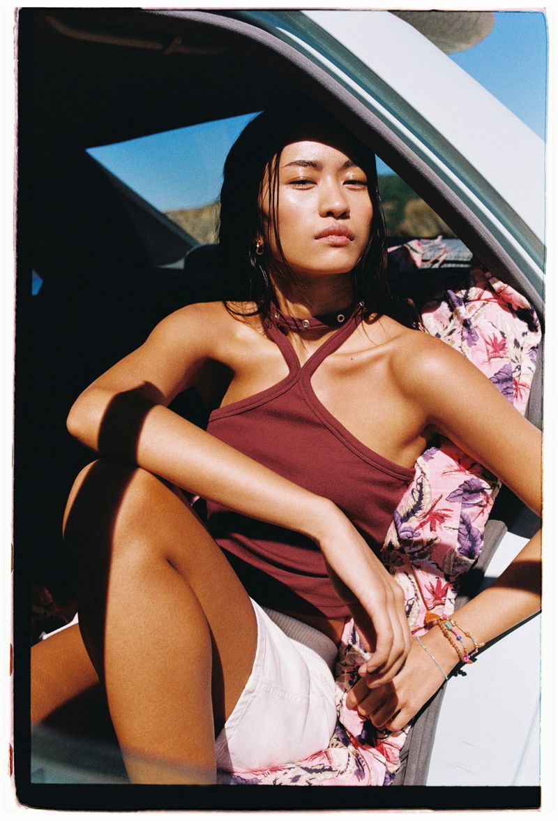Portrait of an Asian woman in a car. She wears a black beanie and a maroon halter top. Outside the car, the sky is clear and sunny.