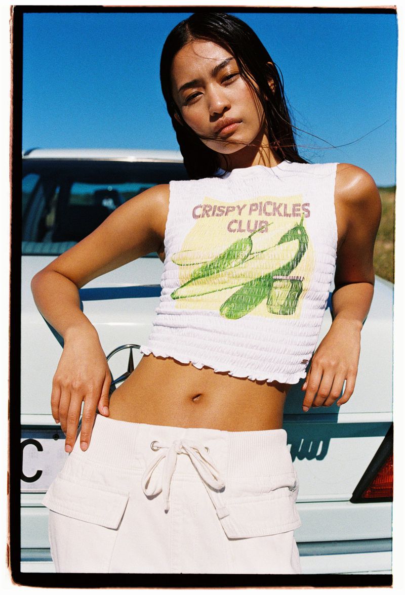 Young Asian woman in a graphic tank top and white skirt poses next to a vintage car against a bright blue sky on a sunny day.