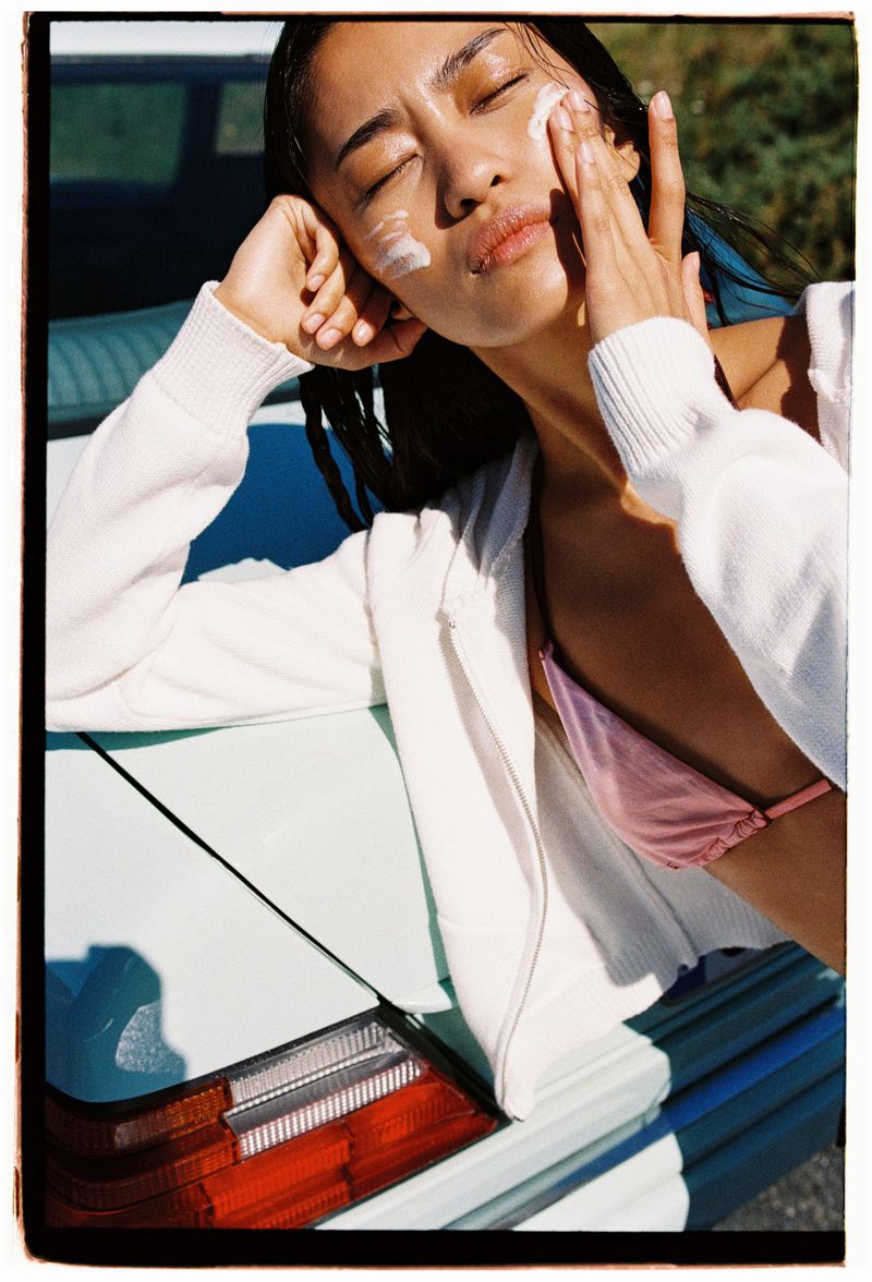Young Asian woman applies sunscreen to her face while leaning on a light green car, creating a vibrant and summery beauty shot.