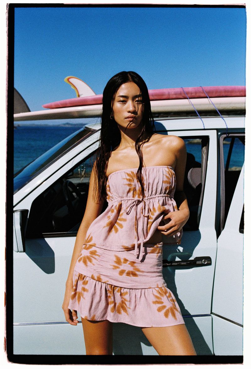 A woman in a strapless dress stands in front of a light blue van with surfboards on top on a sunny day at the beach.