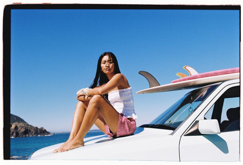 Adult woman in white top and pink shorts sits on a white car with surfboards on top with a blue sky and ocean in the background.
