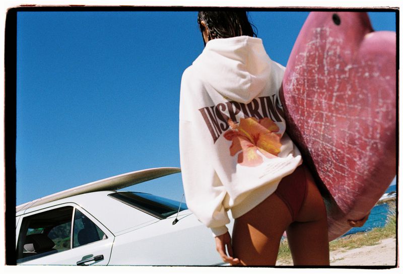 An adult female carrying a surfboard, wearing a white hoodie with a graphic design and car with a surfboard on top in background.
