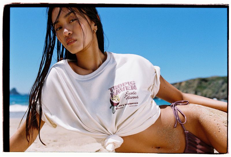 Asian woman poses on beach wearing a white t-shirt and bikini bottoms against blue sky. T-shirt has 'Tropic Haven' logo.