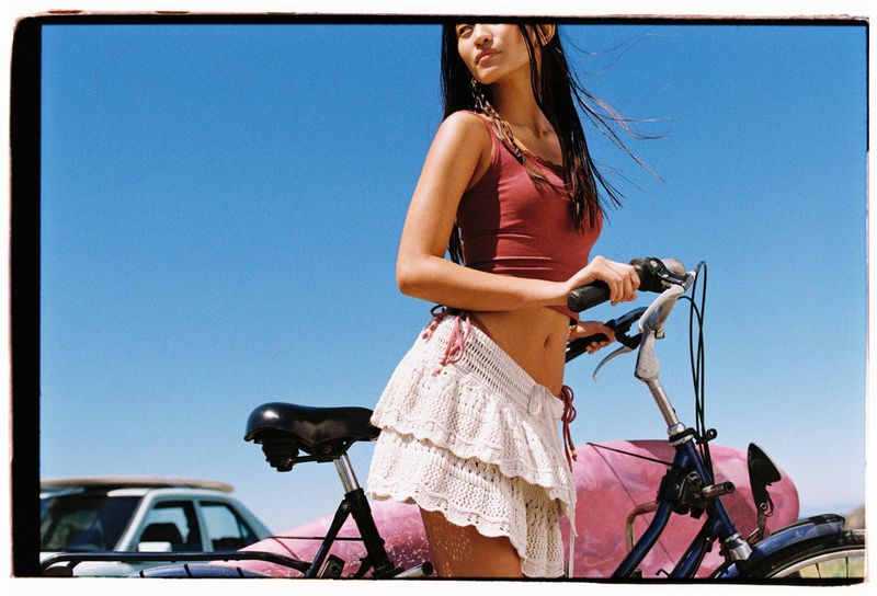 Woman in a red tank top and white skirt with a bicycle at the beach on a sunny day with a blue sky in the background.