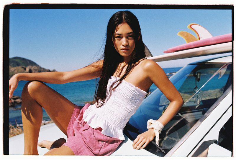 Young woman in pink shorts and white top sits on a car with surfboards at the beach on a sunny day, showcasing a fashion lifestyle shoot.