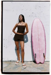 Model standing outdoors next to a pink surfboard wearing a dark brown tube top, matching shorts, and platform sandals.