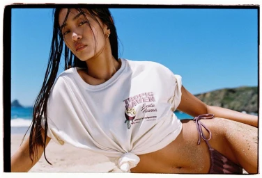Model with damp hair wearing a white graphic t-shirt and bikini bottom reclining on a sandy beach against a bright blue ocean background