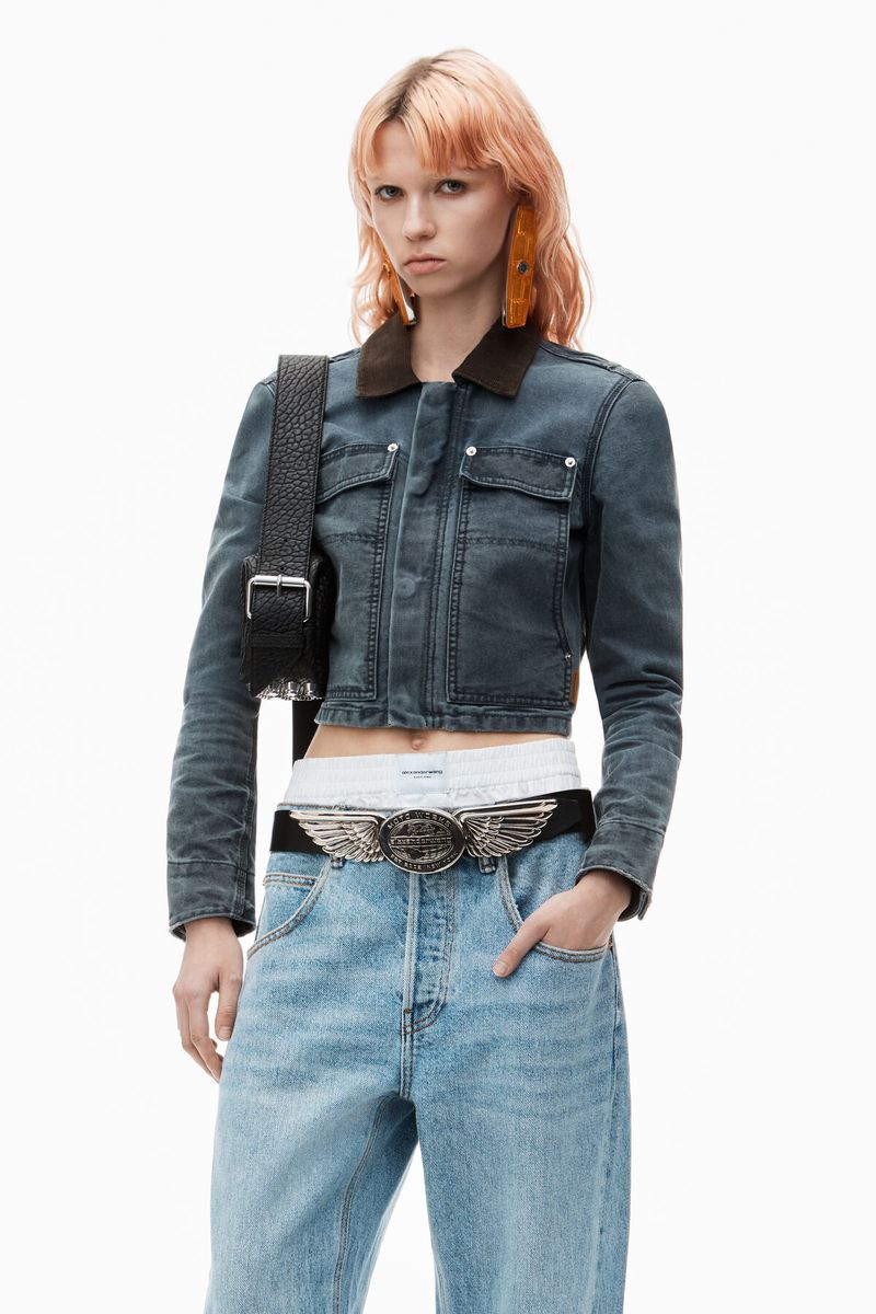 Blonde woman with denim jacket and jeans. She poses in front of a white backdrop, holding her hand in her pocket.