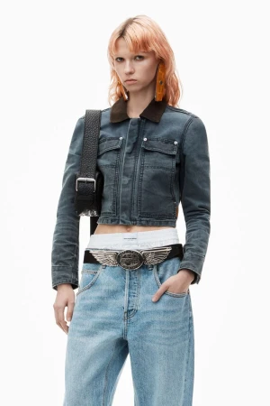 Blonde woman with denim jacket and jeans. She poses in front of a white backdrop, holding her hand in her pocket.