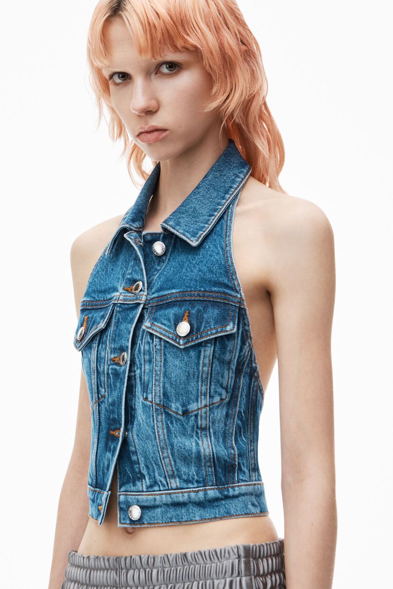 Close-up of a young adult woman with pale skin and pink hair wearing a sleeveless blue denim halter top against a white background.