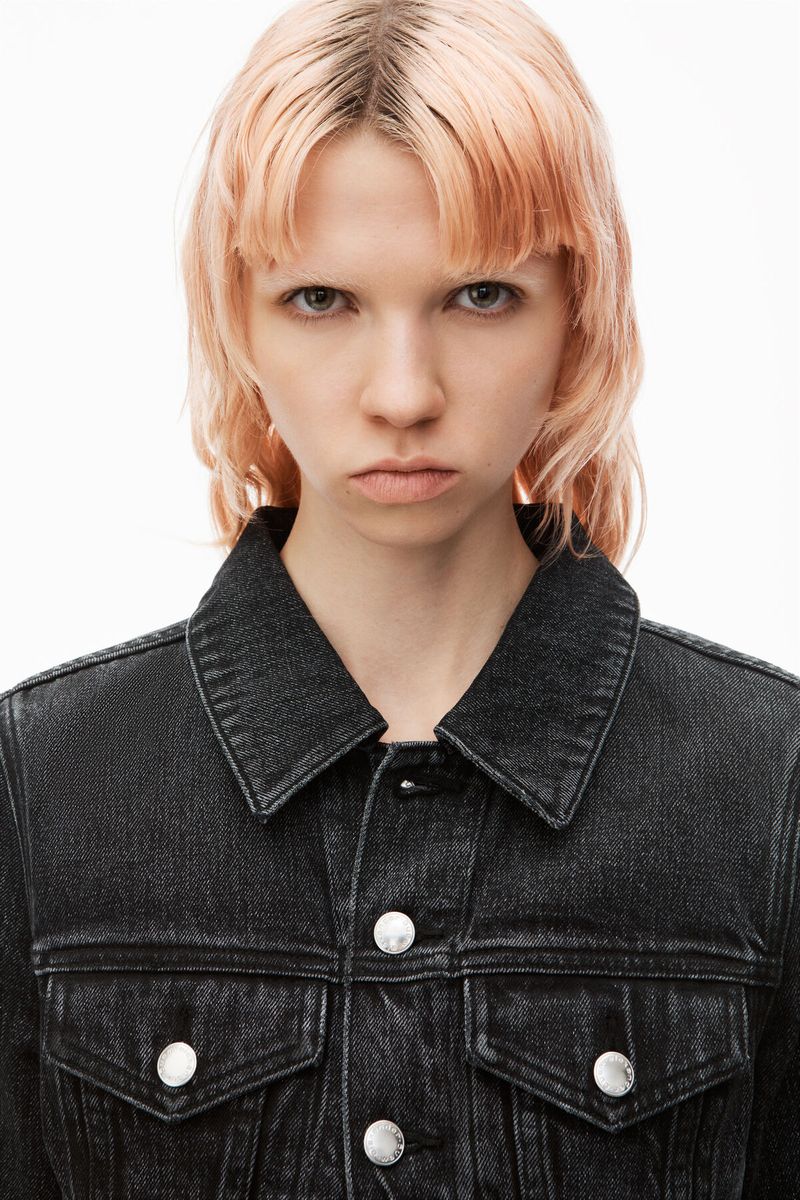 Close-up studio shot of a young adult woman with peach-toned wavy hair, wearing a dark-wash denim jacket and a neutral facial expression.