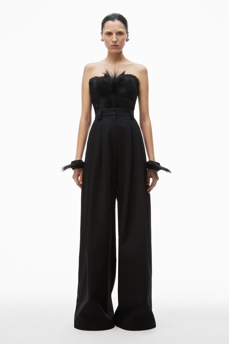 Fashion shot of a female model wearing a black strapless top with feather detail and wide-legged pants, set against a clean white backdrop.