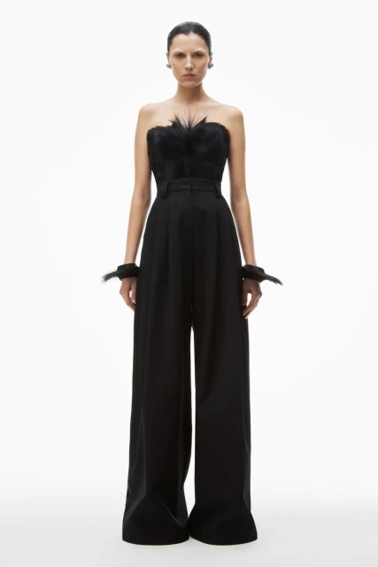 Fashion shot of a female model wearing a black strapless top with feather detail and wide-legged pants, set against a clean white backdrop.