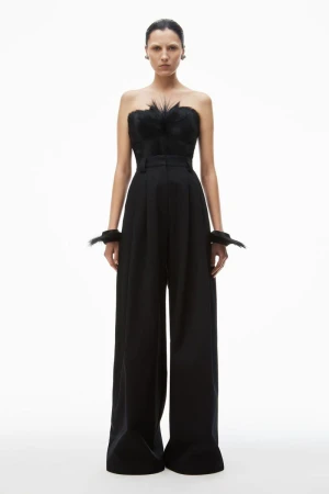 Fashion shot of a female model wearing a black strapless top with feather detail and wide-legged pants, set against a clean white backdrop.