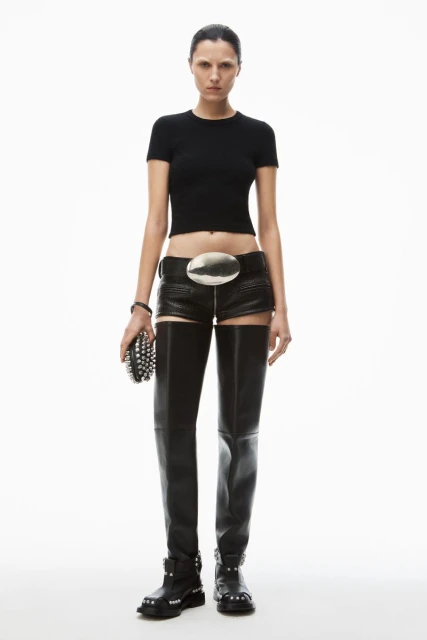 Fashion model strikes a pose in a studio wearing a black top, leather shorts with thigh-high leggings, and carrying a studded clutch.