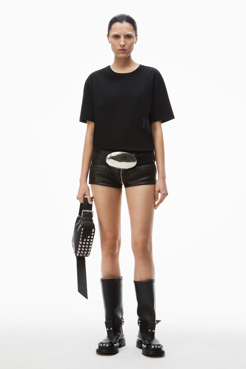 Full-length studio shot of a woman wearing a black t-shirt, black shorts, studded boots, and carrying a studded bag on a white background.