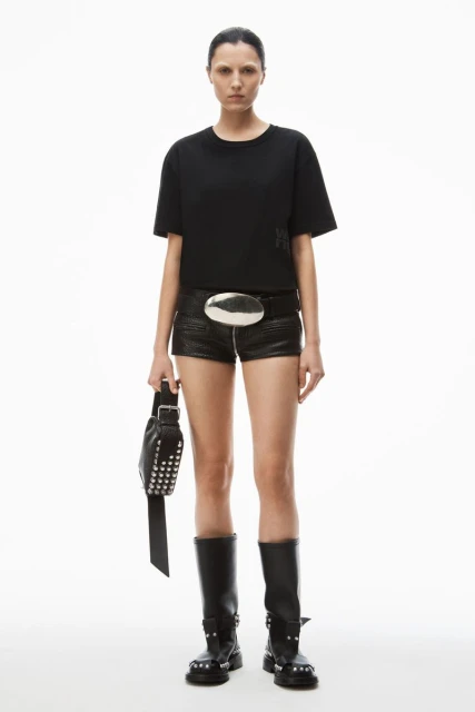 Full-length studio shot of a woman wearing a black t-shirt, black shorts, studded boots, and carrying a studded bag on a white background.