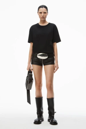 Full-length studio shot of a woman wearing a black t-shirt, black shorts, studded boots, and carrying a studded bag on a white background.