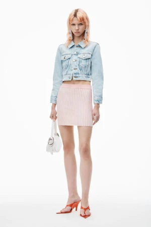 Full-length shot of an adult woman posing in a cropped denim jacket and pink skirt, holding a white purse, against a white backdrop.