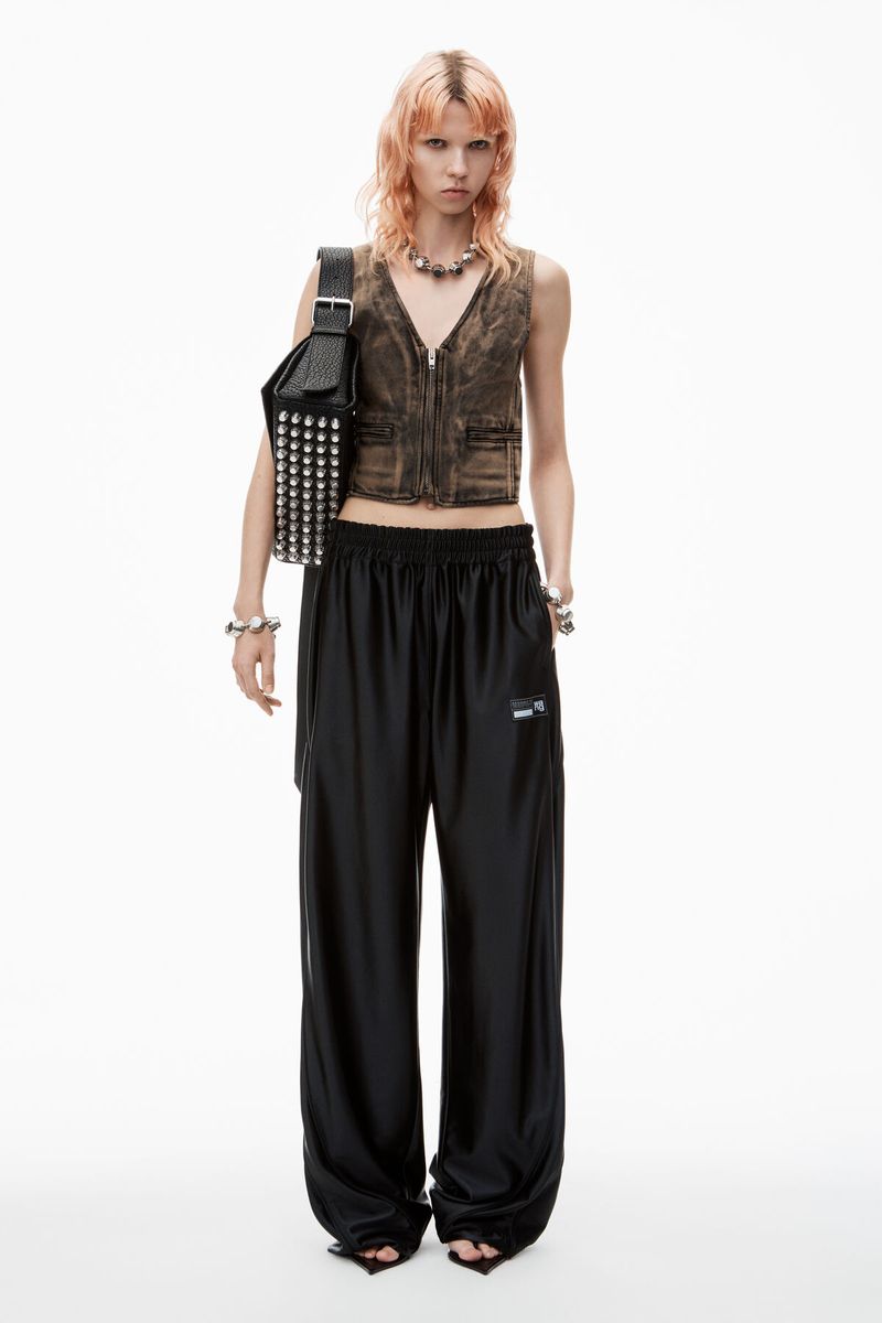 Full-length shot of a woman with pink hair wearing a vest, necklace, bracelet, and black pants against a white background.