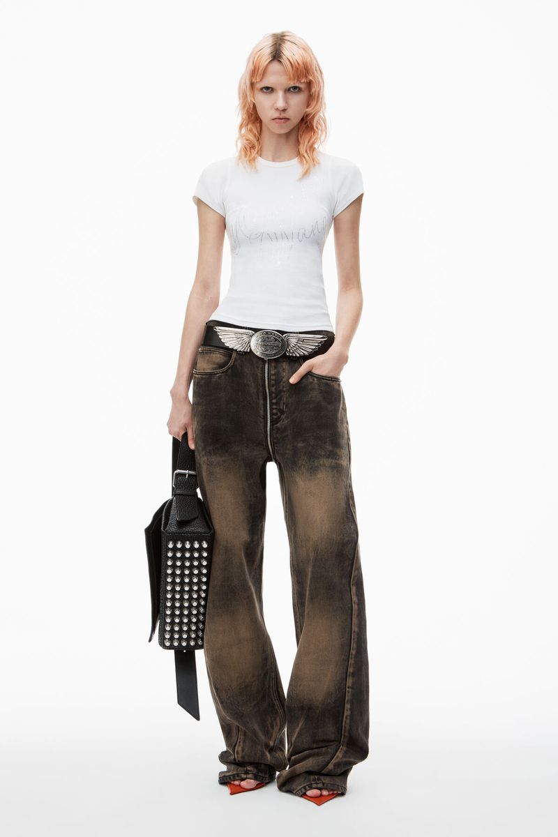 Fashion portrait of a female model in a white top and brown jeans, holding a studded black bag, in a photo studio setting.