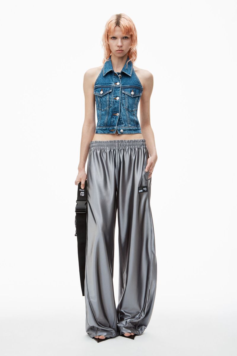 Fashion model poses in a halter-style denim vest and silver wide-legged pants, holding a black crossbody bag against a white background.